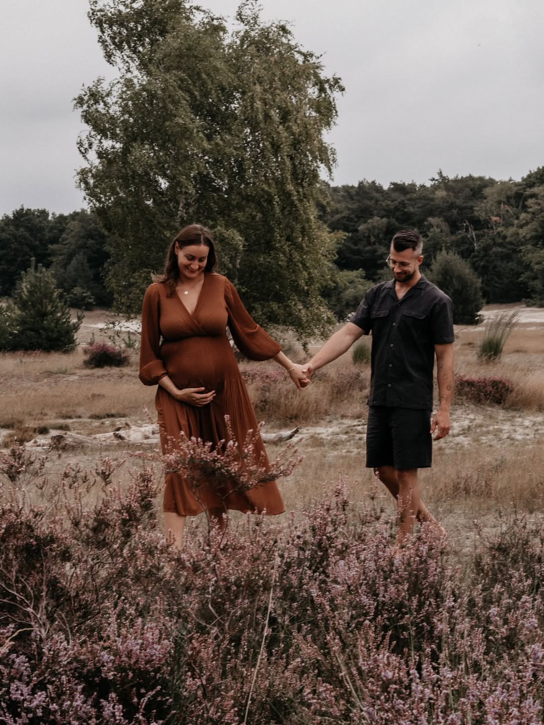 Zwangere vrouw in roestbruine jurk loopt hand in hand met haar partner door de heide